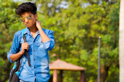 Confident young man standing against trees on sunny day