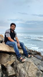 Portrait of smiling man sitting on rock at sea shore against sky