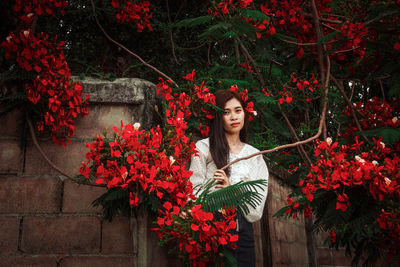 Portrait of young woman standing by red flowering plants