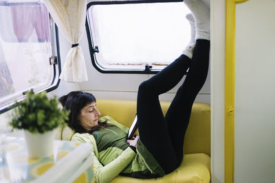Side view of woman lying on sofa reading ebook, legs leaning wall
