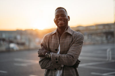 Cheerful man with arms crossed standing at parking lot during sunset