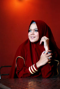 Smiling young woman in hijab looking away while sitting against red wall