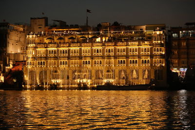 Illuminated buildings by river against sky at sunset