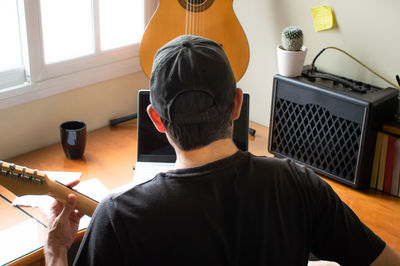 Rear view of man playing guitar while using laptop at home