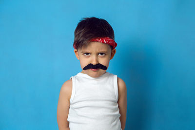 Small boy with a false mustache in a white t-shirt and a red headband on a blue background