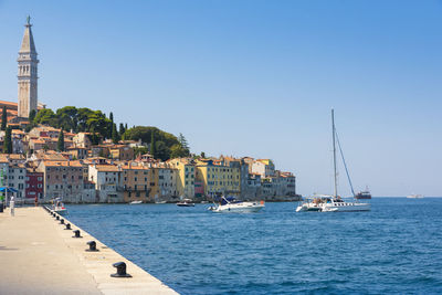 Sailboats in sea by buildings against clear blue sky