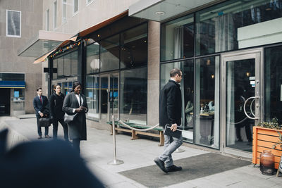 Businessman walking inside store while male and female colleagues standing in line on footpath during covid-19