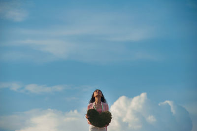 Woman holding heart shape while looking up against sky