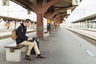 Full length side view of young businessman using laptop on railroad station