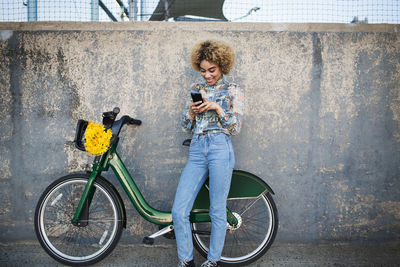 Woman using phone while standing by bicycle against wall