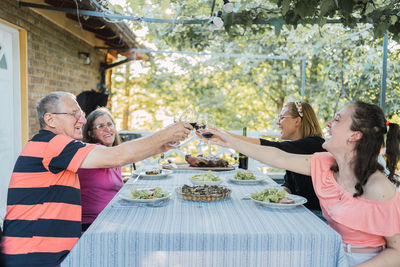 Side view of cheerful family with senior spouses and young daughters smiling and clicking glasses of red wine during family dinner on terrace in countryside