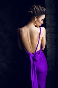 A young woman stands next to the window back in purple long dress