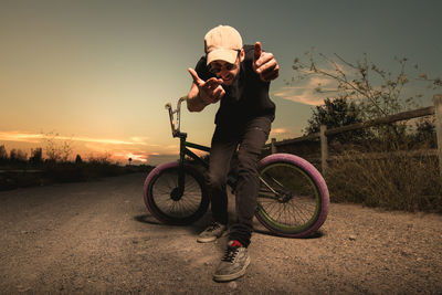 Man with bicycle gesturing against sky during sunset