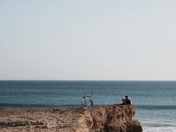 Distant view of man sitting on cliff by sea against clear sky