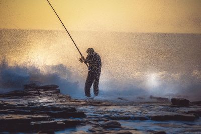 Man fishing in sea against sky during sunset