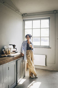 Portrait of smiling female entrepreneur holding disposable cup and leaning on table at workshop