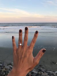 Cropped hand of woman showing wedding ring at beach against sky during sunset