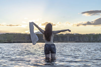 Rear view of woman with arms raised holding top standing in lake against sky