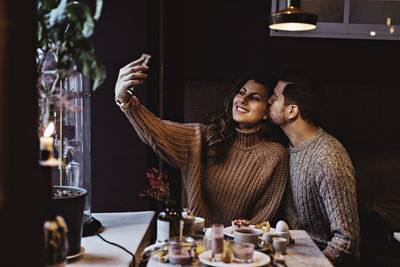 Smiling woman taking selfie through smart phone while male partner kissing in hotel
