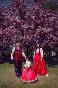 Korean family in national costumes in nature stands next to a cherry blossoming tree.