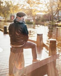 Rear view of thoughtful man sitting on wood by lake at public park