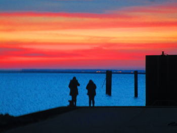 Silhouette friends standing on beach against orange sky