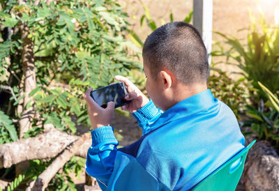 Back view of child boy playing online games with smartphone, sitting outdoor play with phone in hand 