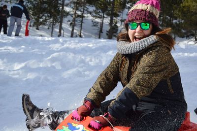 Portrait of happy young woman with sunglasses sitting on sledge during winter