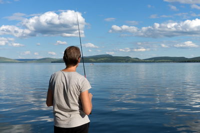 A woman fisherman stands with a fishing rod and catches fish against a blue lake and sky with clouds