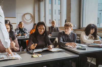 Multiracial students sitting at desk while learning from professor in classroom