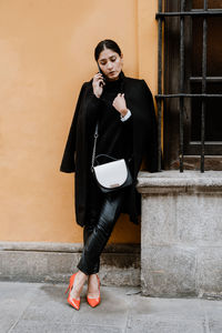 Full body of confident stylish ethnic female entrepreneur with dark hair in elegant black coat and high heels standing near aged building and talking on mobile phone