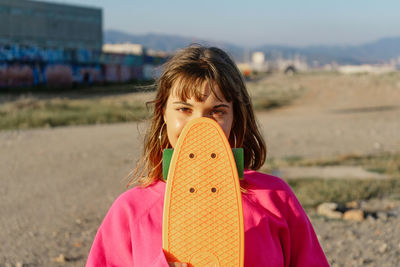 Portrait of young woman covering face with skateboard on land