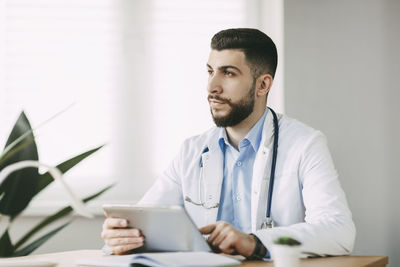 A male doctor of oriental appearance is concentratedly working on a tablet, sitting at a table 