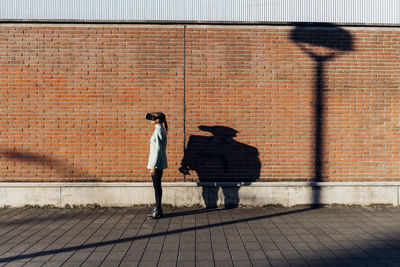 Young businesswoman with vr goggles by wall on sunny day