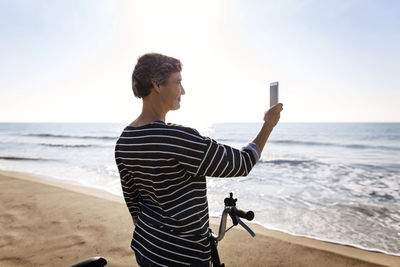Woman photographing through mobile phone while standing with bicycle at beach