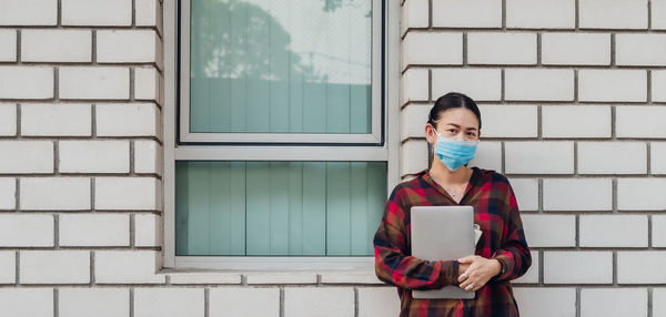 Portrait of woman wearing mask holding laptop standing against wall