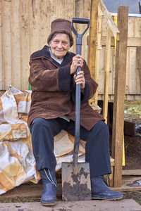 Full length portrait of smiling senior woman with spade sitting in backyard