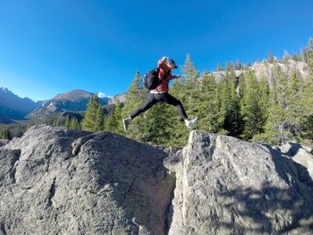 Full length of female hiker jumping on rocks against clear blue sky