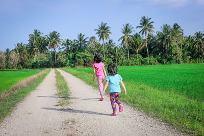 Rear view of sisters walking on dirt road at farm against trees