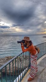 Woman standing on railing by sea against sky during sunset