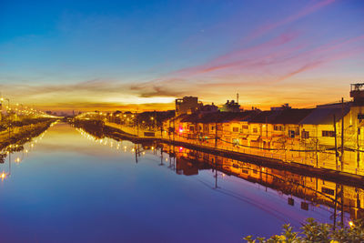 Illuminated houses reflecting on calm river against sky during sunset