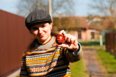 Ukrainian young smiling woman in hat holding one colored red egg on nature background. easter