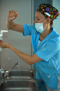 Adult female medic in sterile mask and uniform applying liquid sanitizer on hand above sink at work in hospital