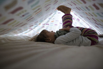 Side view of girl playing while lying underneath blanket at home