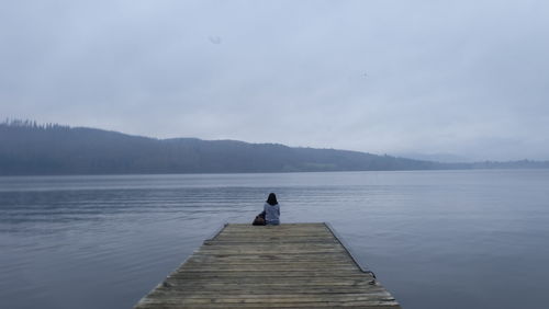 Rear view of woman sitting on pier lake against sky