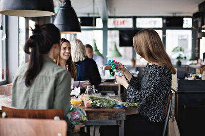 Smiling female friends looking at woman holding gift while sitting at dining table in restaurant