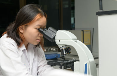 Cute scientist schoolgirl in lab coat looks through microscope for study microbiology in laboratory. 