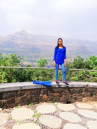 Portrait of woman standing on retaining wall against mountains and sky