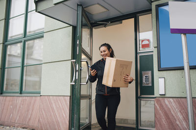 Smiling female worker with box using mobile phone while coming out of building