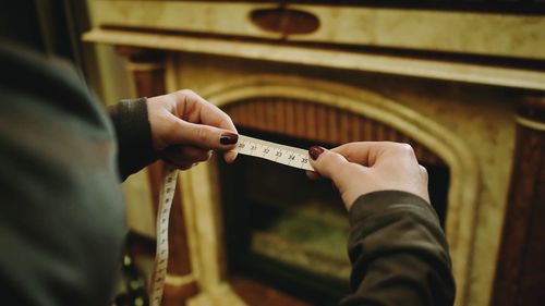 Cropped hands of woman holding tape measure against fireplace